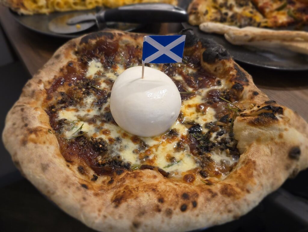 Caramelised onions on a pizza with cheese, haggis, and fresh herbs. Burratta ball sitting on top with a scottish saltire flag on a cocktail stick poked into the burratta.