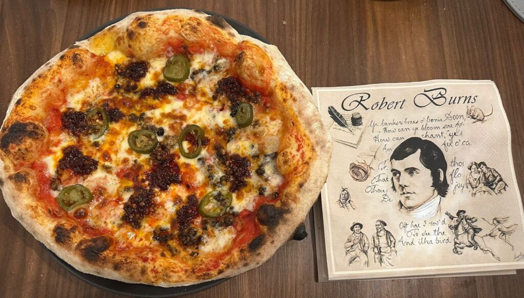 Crumbly haggis, n'duja spicy sauage and jalapenos on a bed of mozzarella on a pizza. With a napkin next to the pizza with Robert Burns's face and poetry extracts