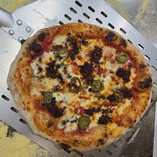 Crumbled Haggis and n'duja on a pizza with sliced jalapenos. All sitting on a pizza peel with some cornmeal on the worktop.